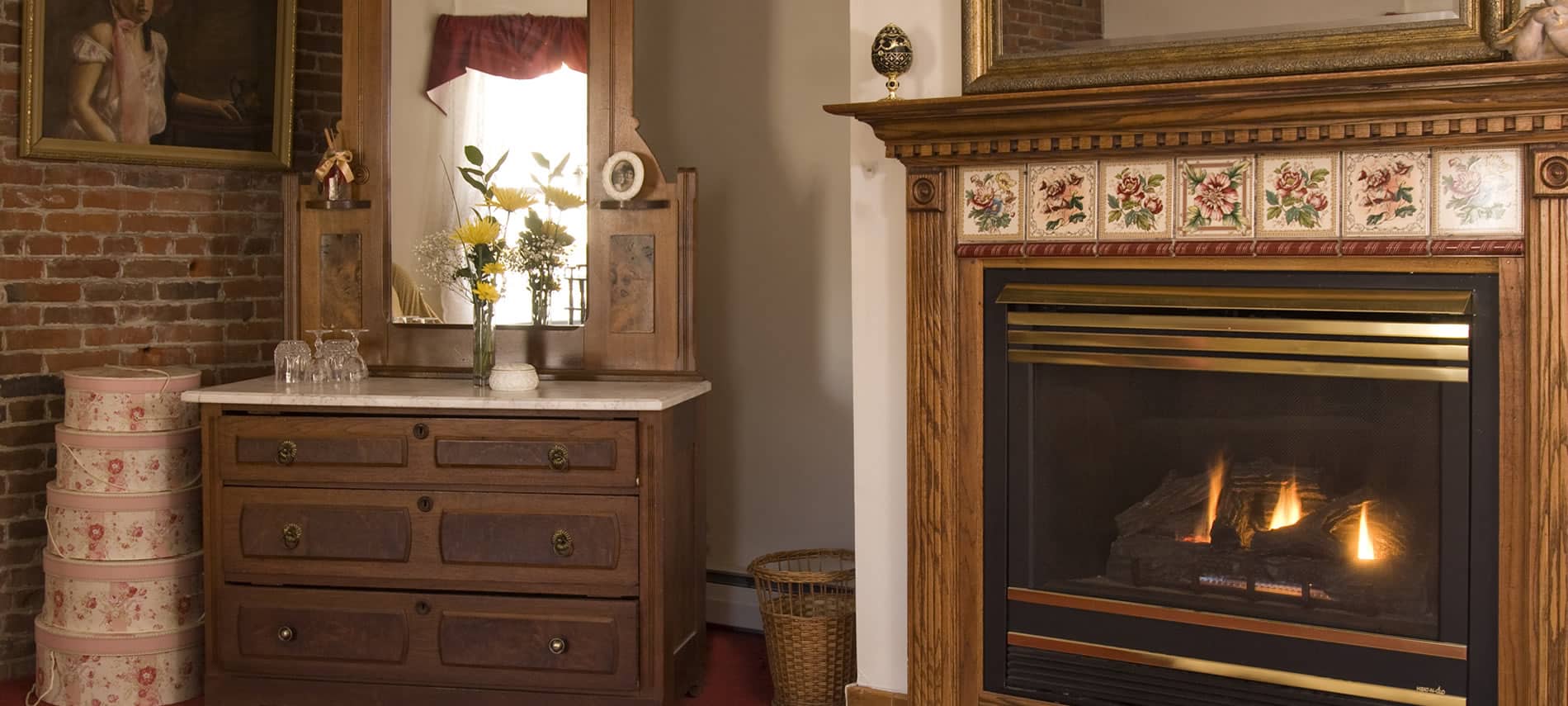 Fireplace with wood wrap around mantel and decorative tile inserts, corner chest with mirror, and brick wall with framed portrait
